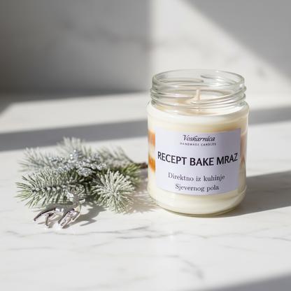 Candle in a glass jar with a label next to a sprig of pine on a light surface.